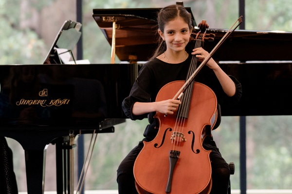 Young girl in black dress and smiling and playing the cello in front of a piano.